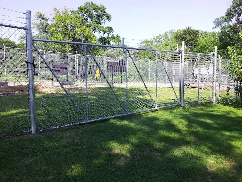 A tall chain-link fence with a double swing gate separates a green lawn from a wooded area under a bright sky.