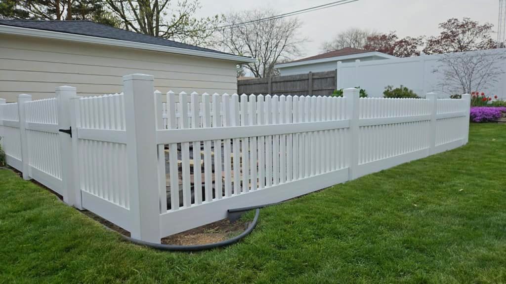 A white vinyl picket fence encloses a backyard lawn, featuring a gated corner entrance next to a house.