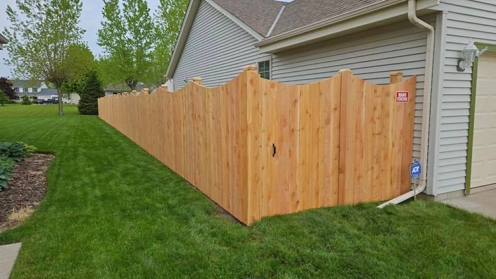 A new, scalloped-top wood privacy fence runs along the side of a house next to a green lawn.