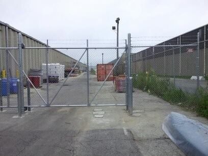A tall metal gate stands closed across a paved industrial passage flanked by high concrete walls and chain-link fencing.