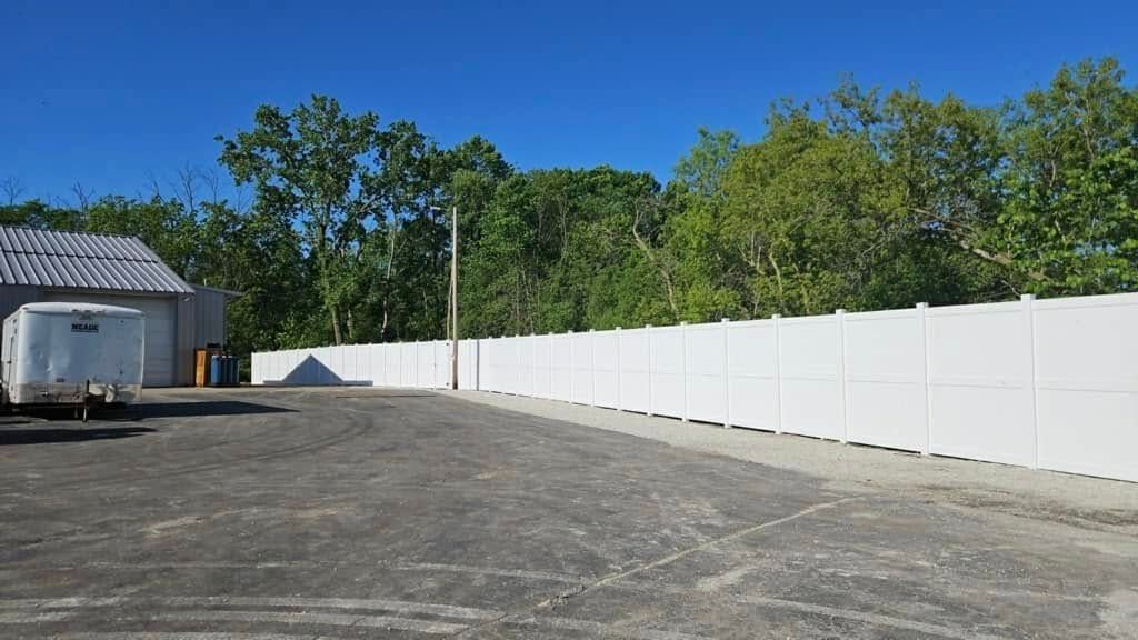 A white vinyl fence borders a paved parking lot next to a building and trees under a clear blue sky.