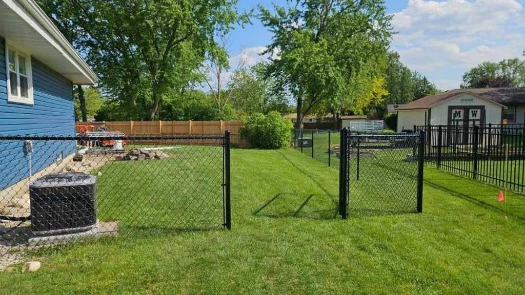 A backyard with a black chain-link fence, a blue house, an air conditioning unit, and a storage shed under a sunny sky.