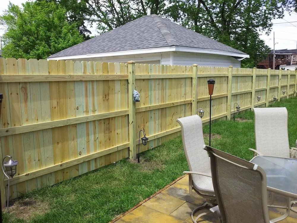 A newly installed light-colored wood privacy fence stands along a grassy lawn next to a backyard patio with chairs.