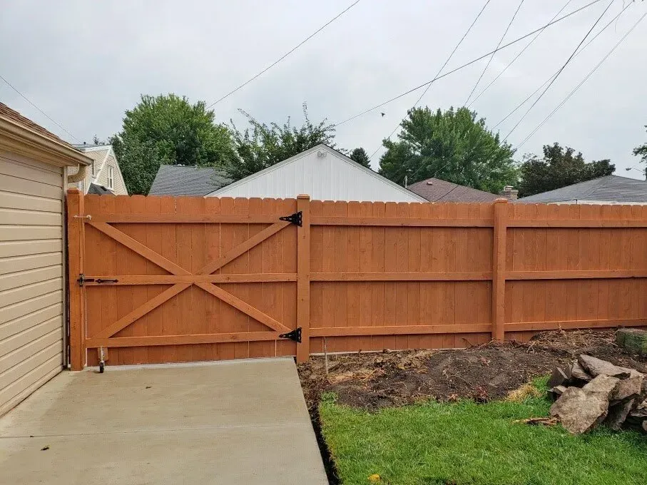 A wooden privacy fence with a cross-braced gate stands next to a house exterior and a concrete driveway.
