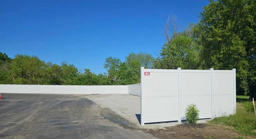 A white vinyl fence borders a paved parking lot next to a line of trees under a clear blue sky.