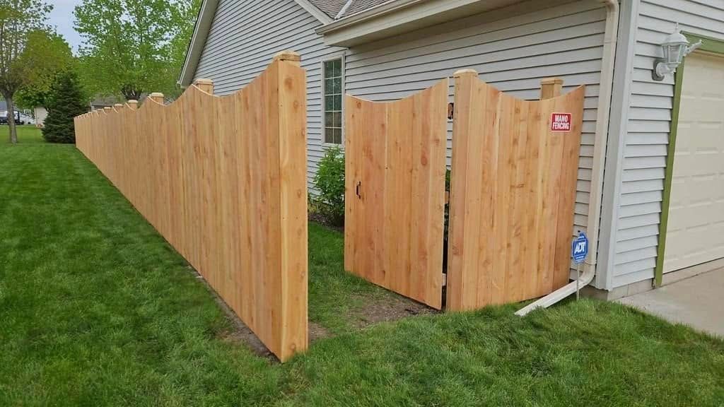 A side view of a new, natural wood privacy fence with a wavy top design, running alongside a house with beige siding.