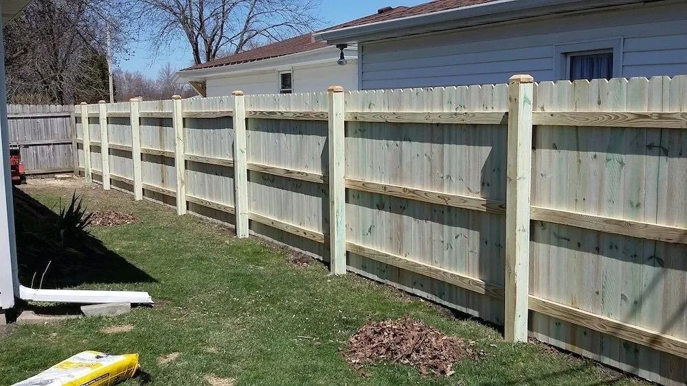 A newly constructed light-colored wooden privacy fence running along a grassy backyard, adjacent to a house.
