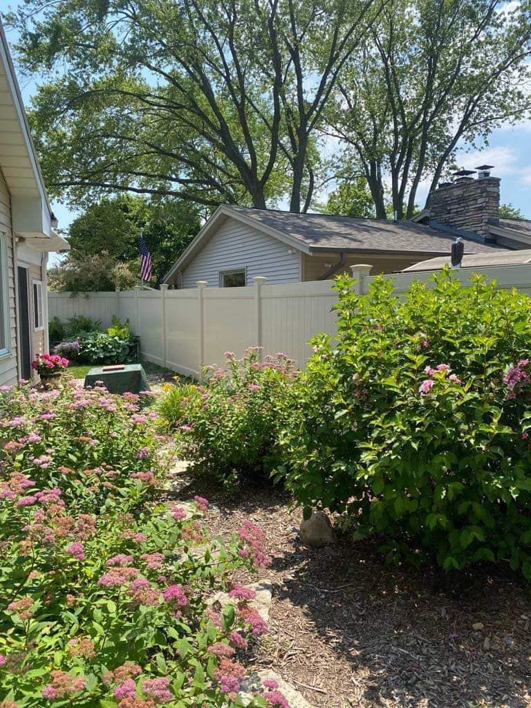 A sunny backyard garden featuring a vinyl fence, lush green bushes with pink flowers, and a house in the background.