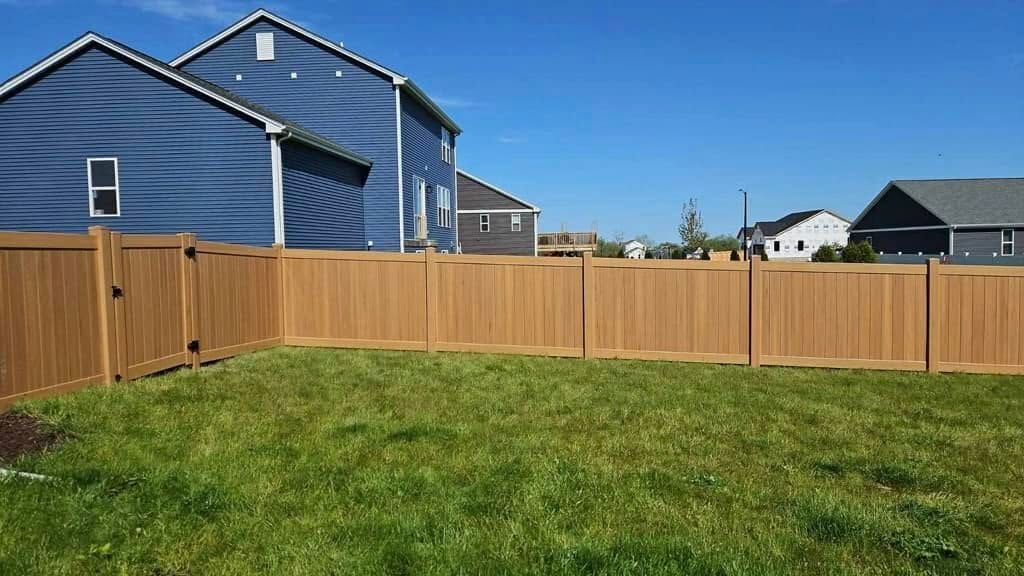 A brown wooden privacy fence enclosing a grassy backyard against a background of suburban houses under a blue sky.