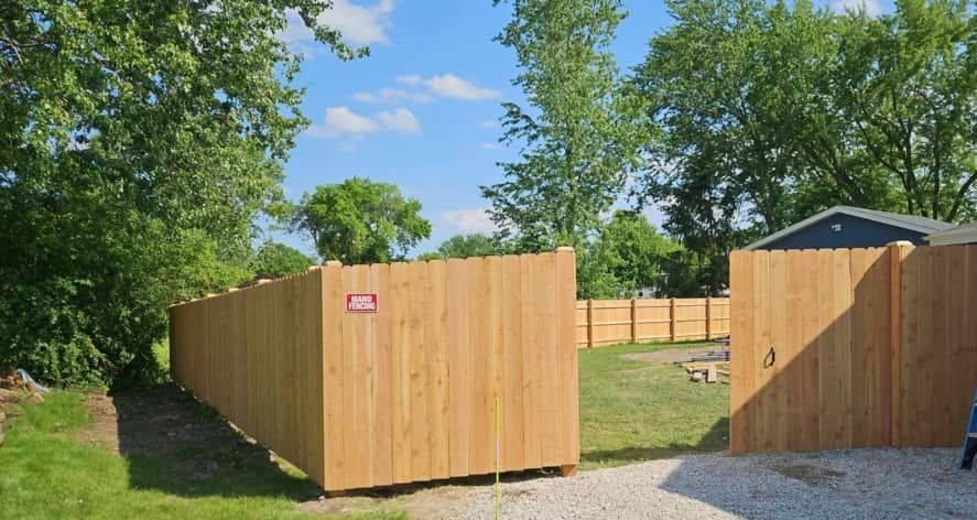 A wooden privacy fence surrounds a grassy backyard area on a sunny day, with a gate opening onto a gravel driveway.