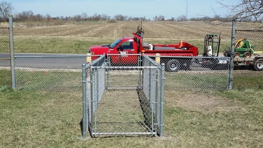 A narrow, chain-link fenced walkway leads to a gap in a perimeter fence, with a red truck parked on a road behind it.