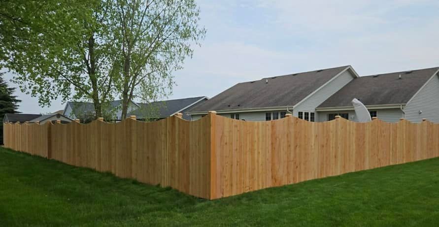 A cedar privacy fence with a wavy scalloped top edge, bordering a residential yard with houses in the background.
