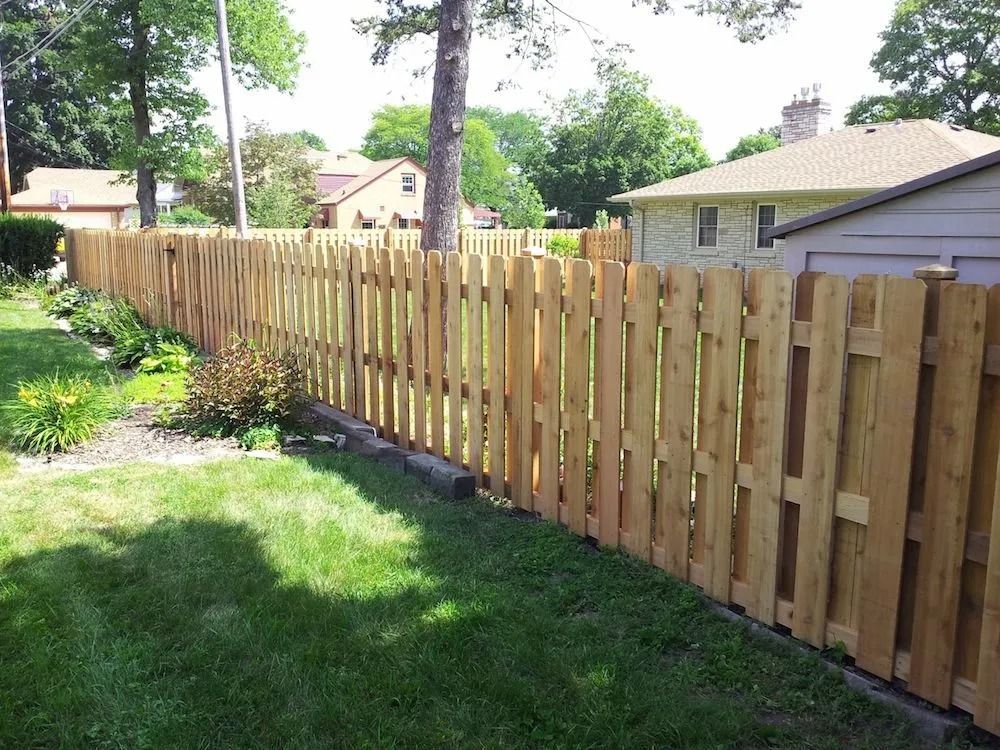 A wood picket fence separates a lush green lawn from neighboring houses on a sunny day.