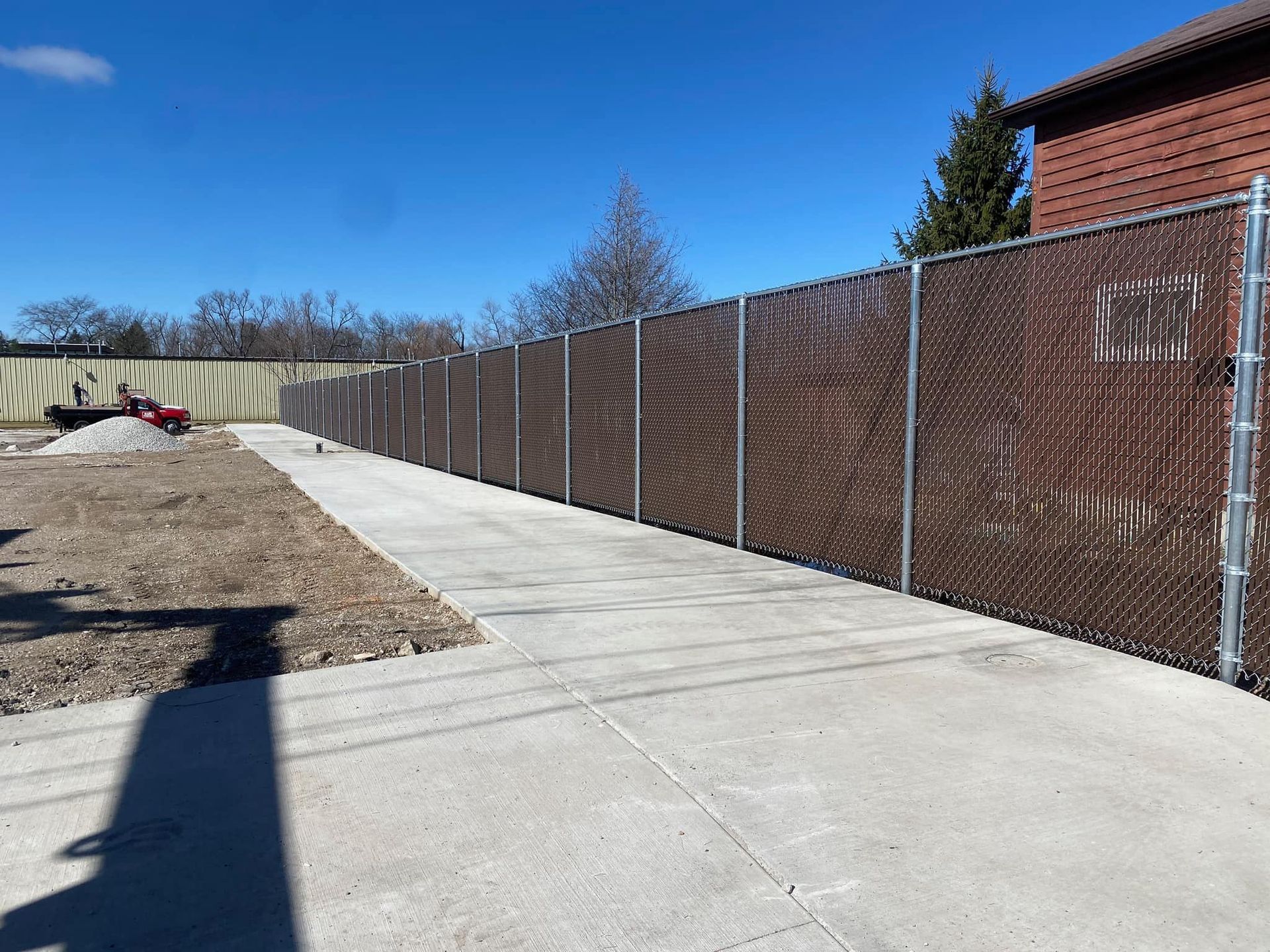 A long chain-link fence with brown privacy slats runs along a concrete sidewalk next to a construction site.
