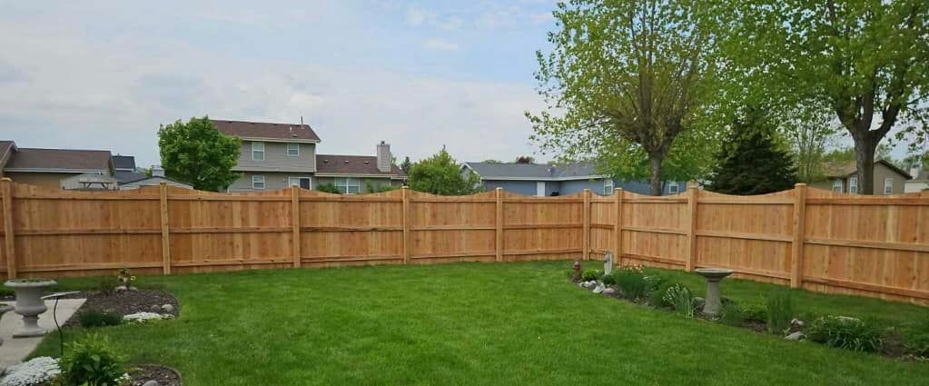 A grassy backyard features a tall, light-brown wood fence, with nearby houses and green trees visible in the background.