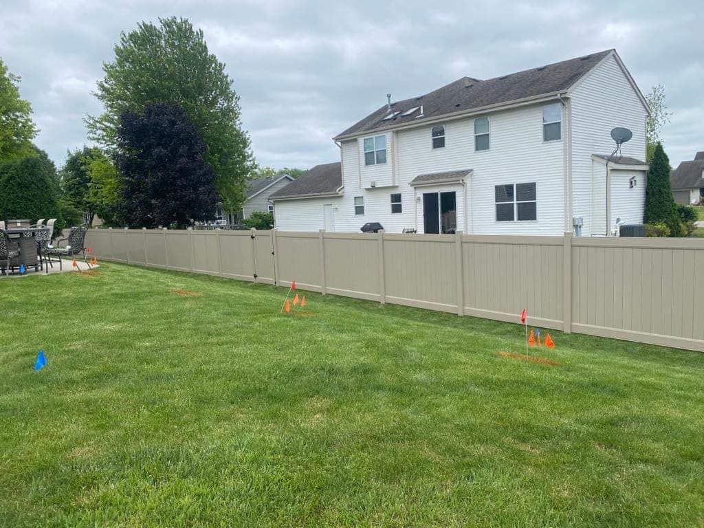 A tan vinyl fence spans a grassy backyard, with small orange utility flags marking the ground in front of a white house.