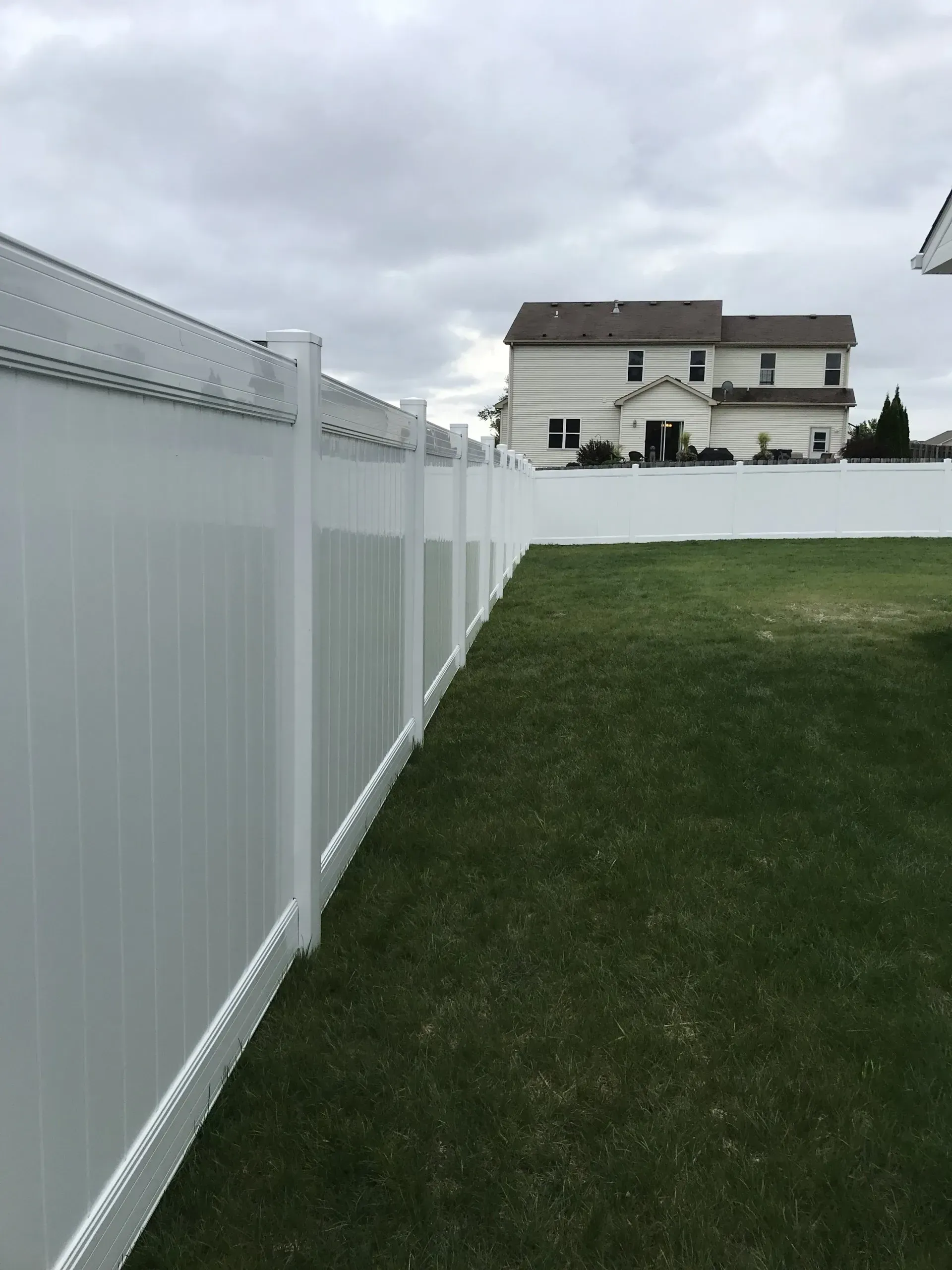 A white vinyl privacy fence runs alongside a green grassy lawn, with a suburban house visible in the background.