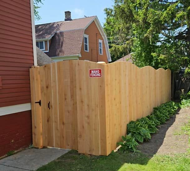 A scalloped-top cedar wood fence and matching gate installed next to a red-sided house with a small garden bed.