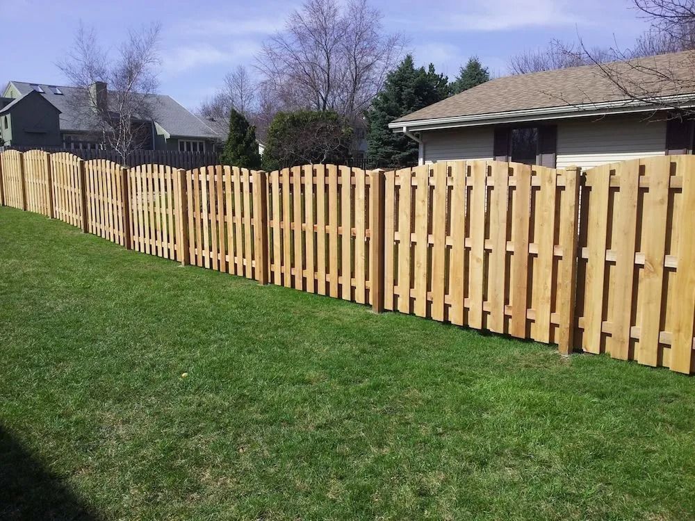 A wooden picket fence with a scalloped top runs across a grassy lawn in front of a house under a blue sky.