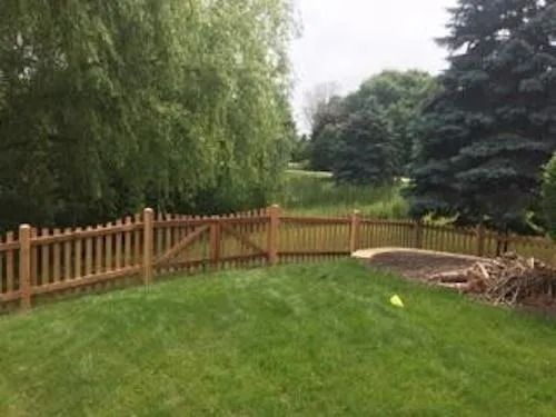 A wooden picket fence borders a green grassy lawn with large trees in the background on a cloudy day.