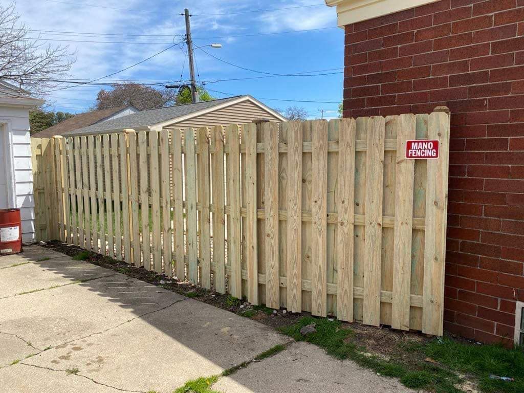 A newly built, light-wood vertical slat fence stands on a concrete driveway next to a brick wall.