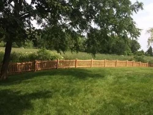 A wooden picket fence stretches across a lush green lawn, bordered by trees and a meadow in the background.