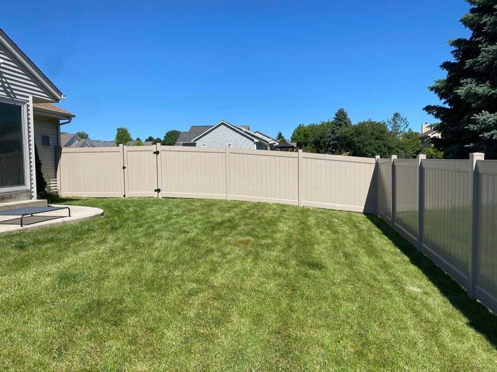 A green backyard lawn bordered by a beige vinyl privacy fence under a clear blue sky.