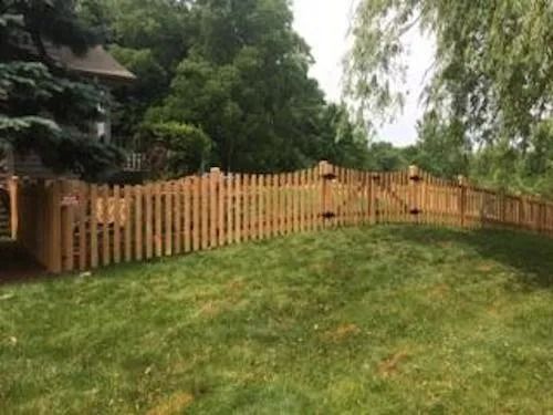 A wooden picket fence with a gate stands in a green yard, with mature trees and a house visible in the background.