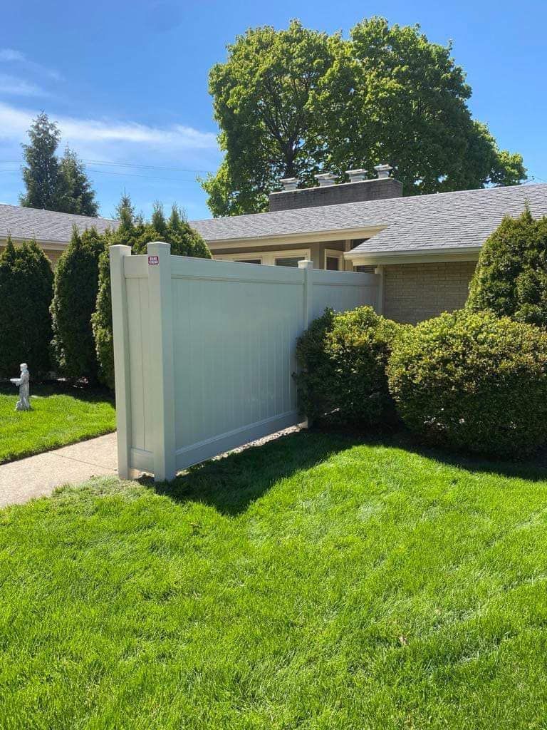 A white vinyl fence stands on a green lawn in front of a house, flanked by manicured green shrubs under a blue sky.