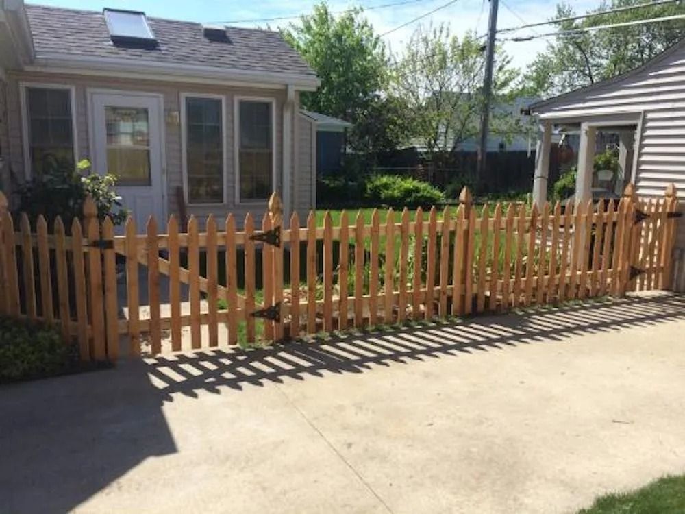 A wooden picket fence with a gate encloses a lawn between two houses on a sunny day.