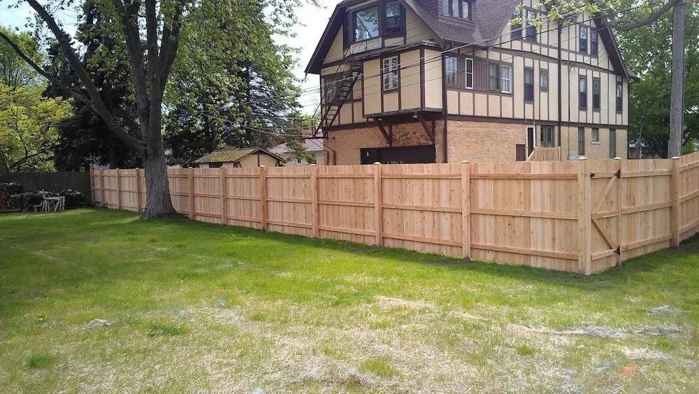 A newly installed tall, light-wood privacy fence surrounding a grassy backyard with a large house in the background.