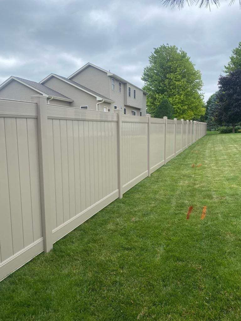 A tall, tan vinyl privacy fence borders a green grassy yard under a cloudy sky.