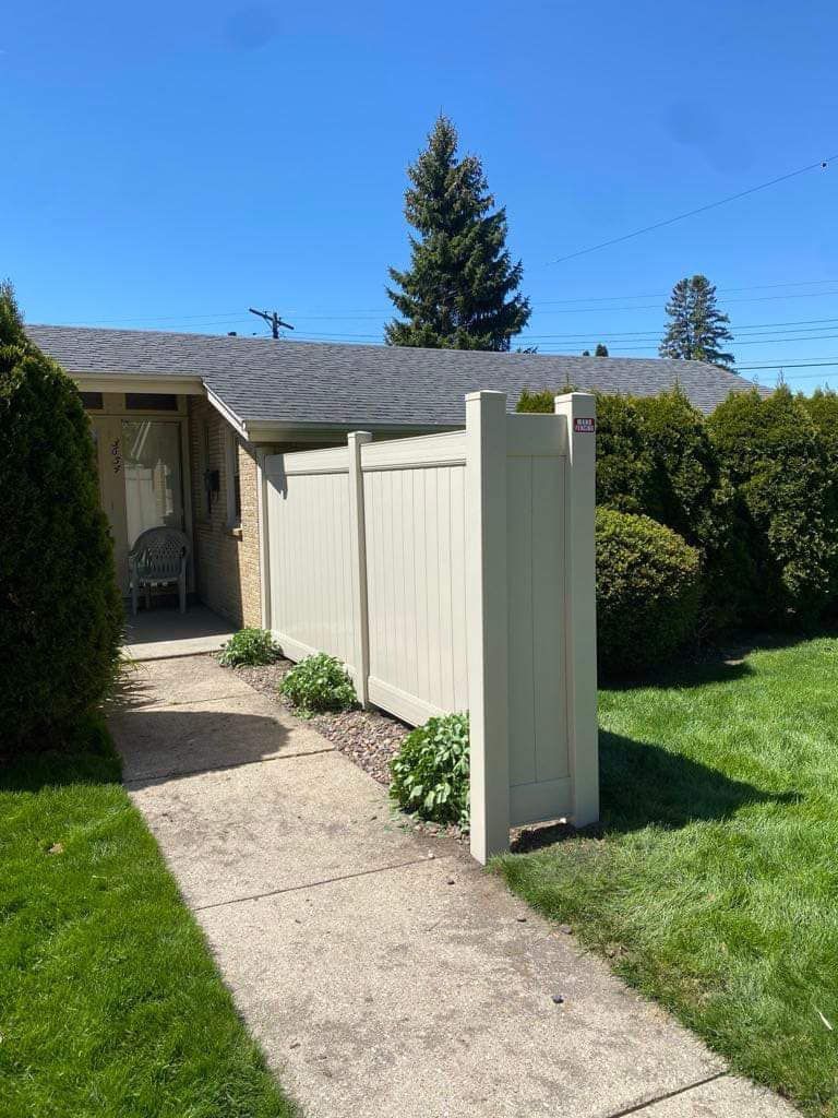 A beige vinyl fence extends from a house, running parallel to a concrete walkway in a green yard under a clear blue sky.