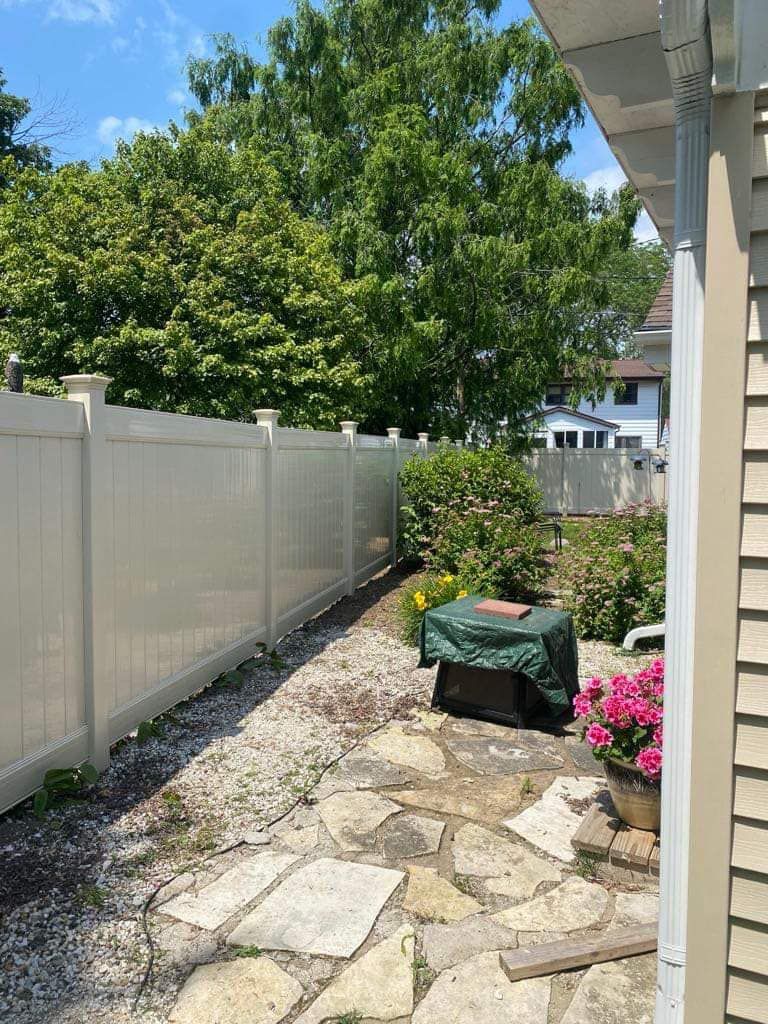 A stone patio bordered by a white privacy fence, a covered grill, and pink flowers against a lush green backyard.