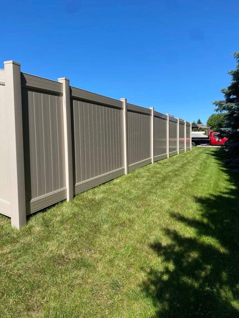 A long, tan vinyl privacy fence stretches across a lush green lawn under a clear blue sky.