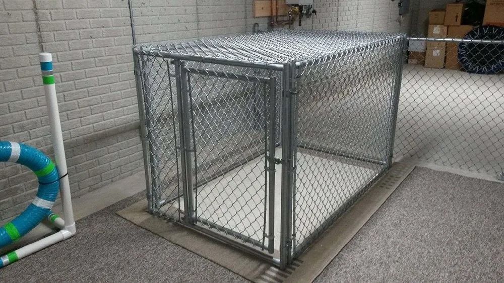 A metal chain-link dog kennel sitting on a concrete floor indoors, next to an agility training tunnel.