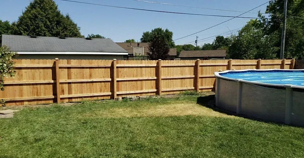 A wooden privacy fence borders a green grassy backyard with a round, above-ground swimming pool under a clear blue sky.