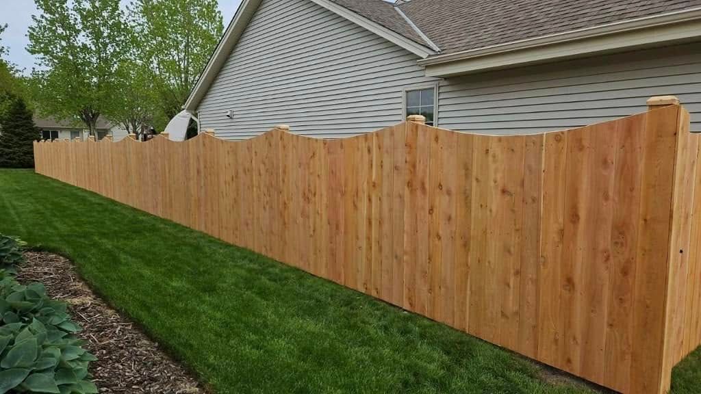 A new, natural-finish wood fence with a wavy, decorative top edge lines a suburban lawn and gray siding house.