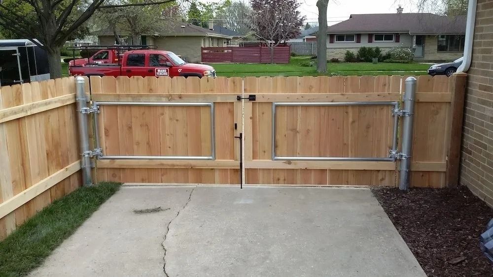A double wooden gate with metal reinforcement frames set in a privacy fence on a concrete driveway.