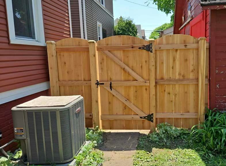A wooden arched gate sits between two houses, next to a gray air conditioning unit on a gravel path.
