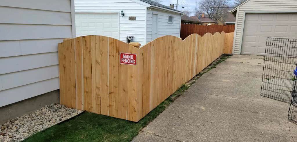 A new, arched-top wooden fence runs along a paved driveway next to a white house.