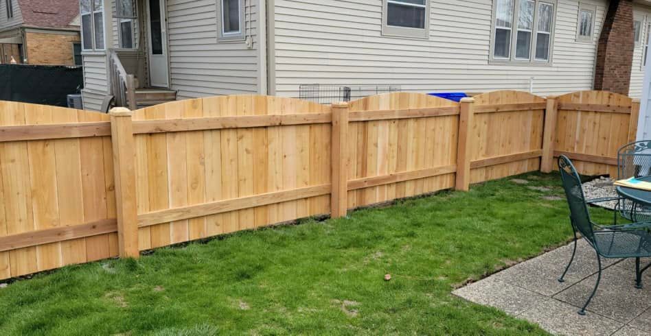 A new, light-brown wood privacy fence with a scalloped top design stands in a grassy backyard next to a house.