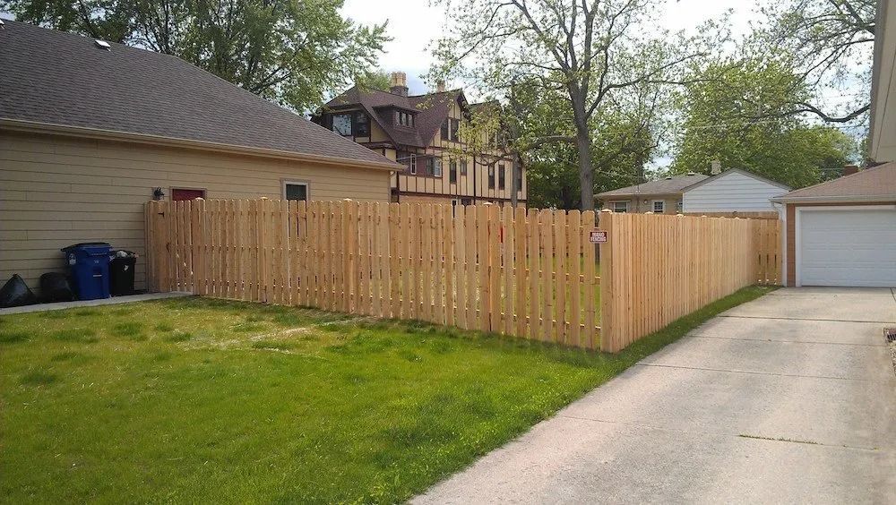 A newly installed wooden picket fence borders a residential backyard next to a driveway and garage.
