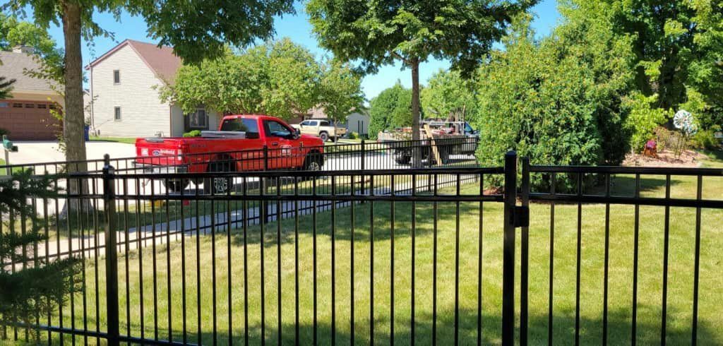 A red pickup truck is parked on a residential street seen through a black metal fence on a sunny day.