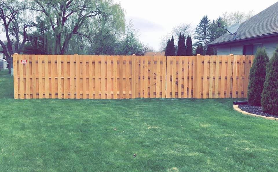 A wooden privacy fence stretches across a green lawn next to a house with evergreen trees.