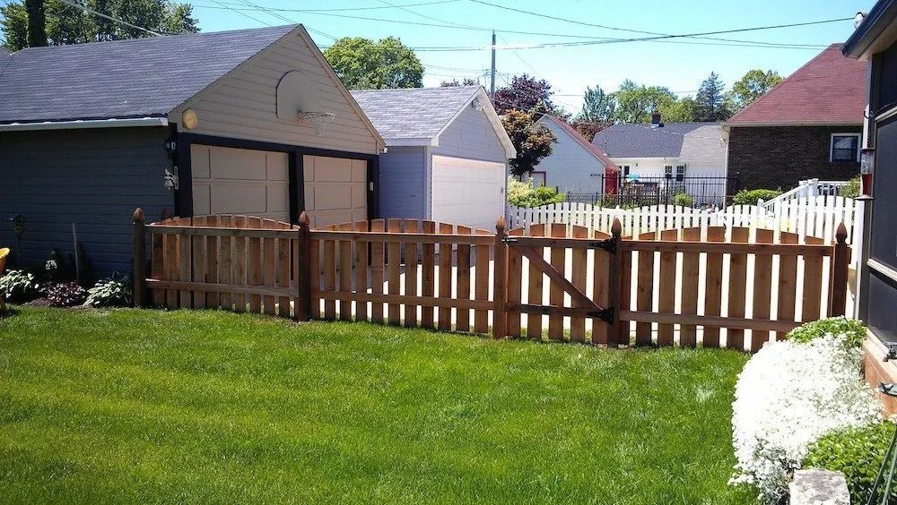 A brown wooden picket fence with a gate encloses a grassy backyard in front of two garages on a sunny day.