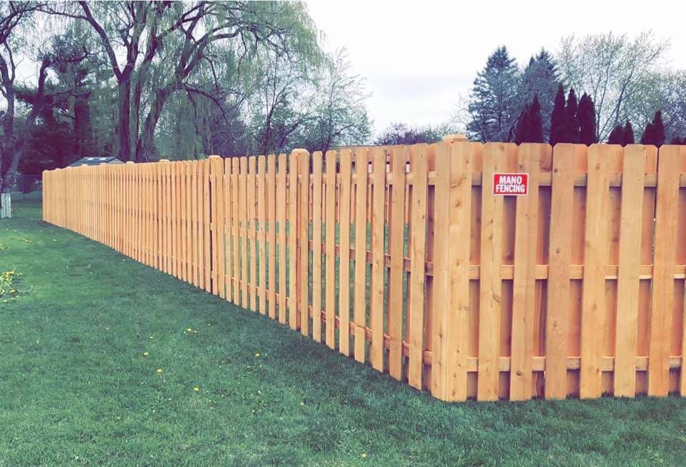 A tall, light-colored wooden picket fence extends across a green grassy lawn under a cloudy sky.