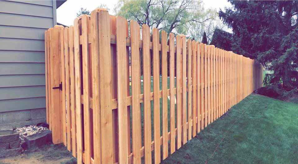A newly installed, light-colored wooden picket fence running along the side of a house and across a grassy yard.