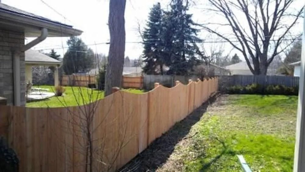A backyard featuring a new, light-colored wooden fence with a decorative scalloped top, built alongside a green lawn.