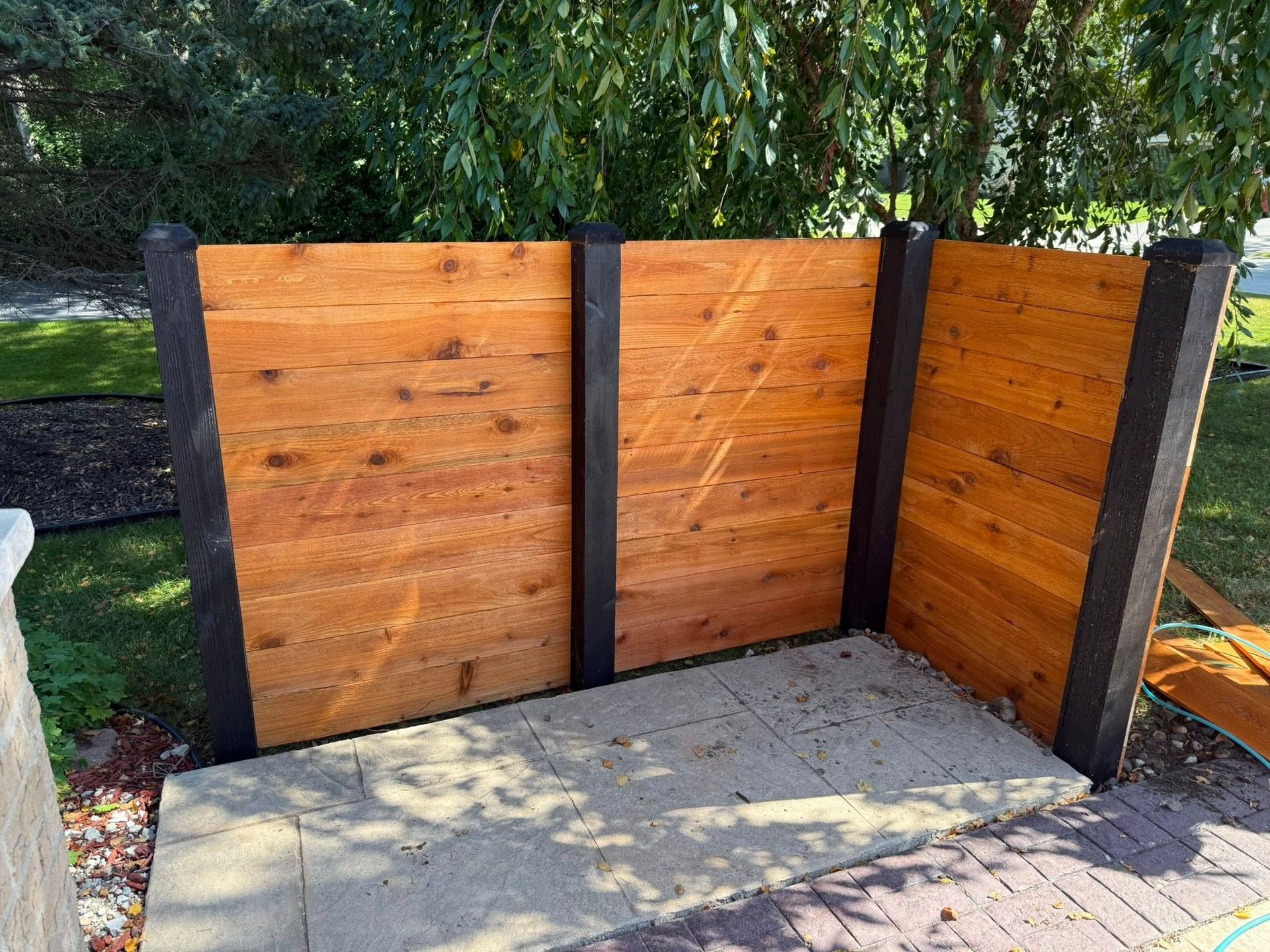An outdoor enclosure made of horizontal cedar wood planks attached to dark, vertical posts on a paved patio.